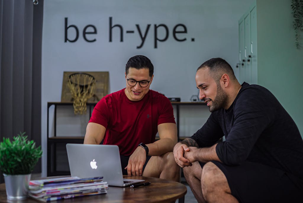 Positive male in eyeglasses sitting and showing information on laptop to smiling bearded man in office
